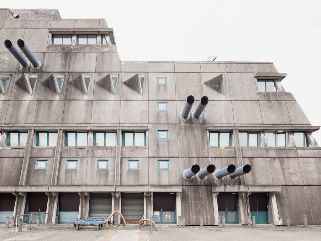 Aus der grauen Zementfassade ragen blaue Lüftungsrohre wie Kanonen aus einem Kriegsschiff heraus