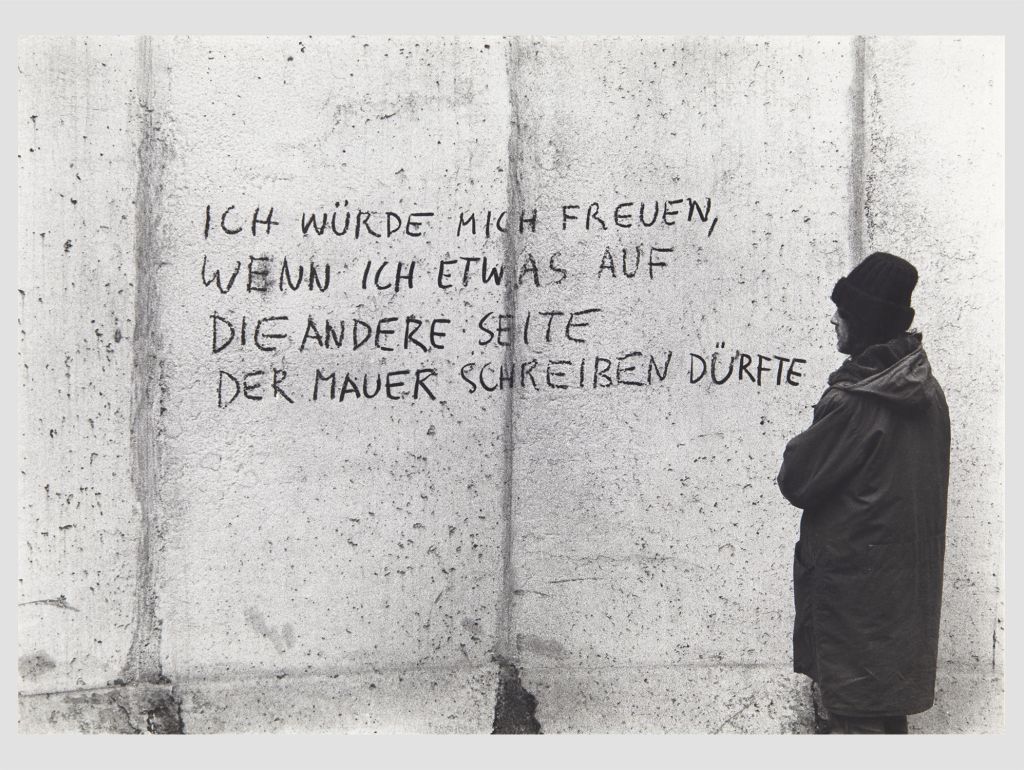 Ein Mann läuft an der mauer in berlin vorbei. darauf steht: ich würde mich freuen wenn ich etwas auf die andere seite der mauer schreiben dürfte