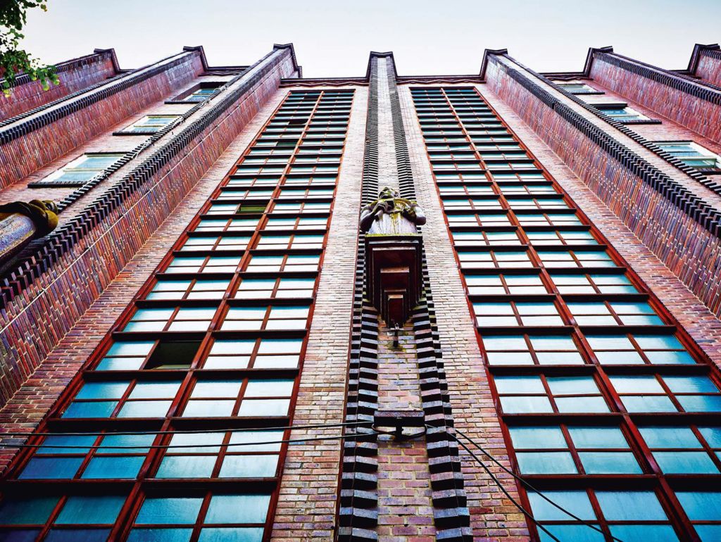 Eine symmetrische Fassade mit großen, vertikalen, rot gerahmten Fenstern gesehen von Unten nach Oben. In der Mitte der Fassade eine Bronzefigur von Henrich Zille. 