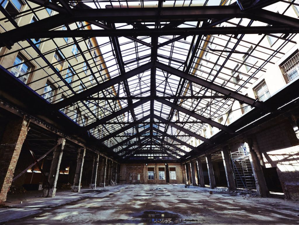 Weite Innenaufnahme eines geräumigen, leeren Lagerhauses mit einem hohen Glasdach. Die Wände bestehen aus freiliegenden Ziegeln und Metallträgern, und Sonnenlicht fällt durch die Metallträgern und wirft Schatten auf den Boden.