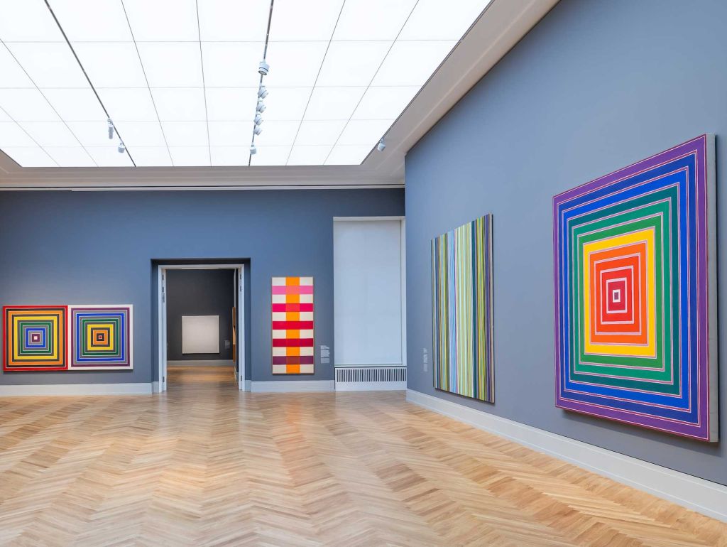Ausstellungsansicht blaue Wände, helle deckenlichter bunte Gemälde an der Wand