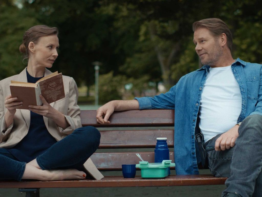 Ein Mann und eine Frau auf einer Parkbank, sie ist barfuss und liest er schaut sie an. Zwischen ihnen Tupperdosen