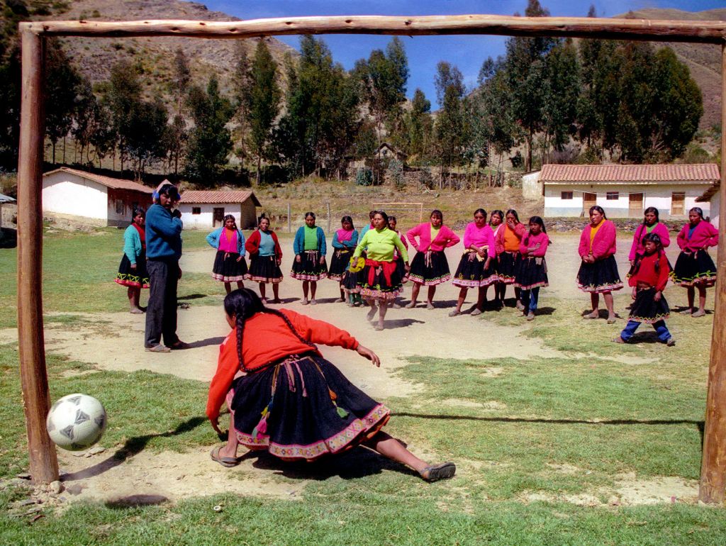 Frauen spielen Fußball im ruralen Kontext. Sie tragen bunten Klamotten. Das Foto ist von der Perspektive der Torwartin aufgenommen.