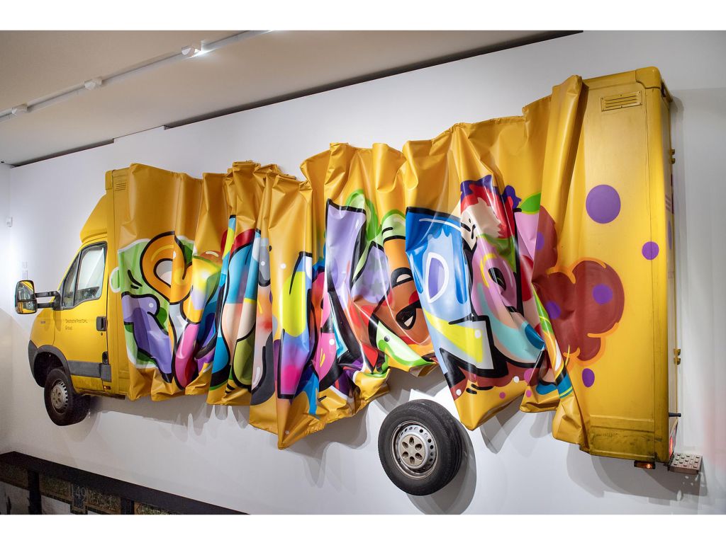 ein halbes Postauto an der wand, zusammengeknautscht und besprüht mit einem Graffitto