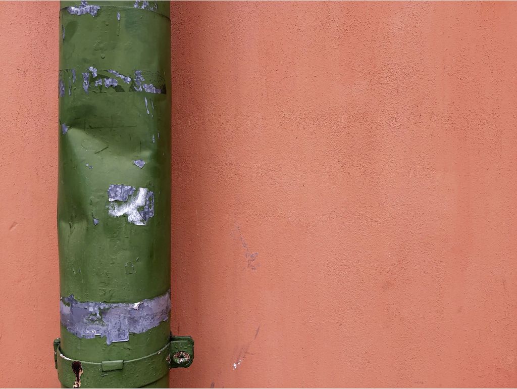 Grünes, verwittertes Fallrohr vor einer orangefarbenen Hauswand, dessen Farbe an mehreren Stellen abgeblättert ist.