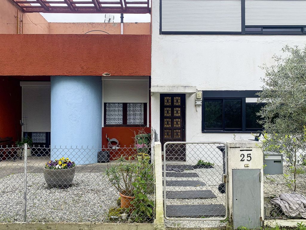 Verschiedenfarbige Hausfassaden mit blauem und terrakottafarbenem Anstrich, davor ein Kiesgarten mit Pflanzen und Maschendrahtzaun.