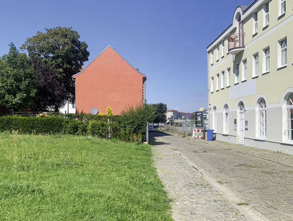 Straßenszene mit einem orangefarbenen Haus und einem hellgrünen Gebäude mit weißen Fenstern; dazwischen ein gepflasterter Weg und Grünflächen.