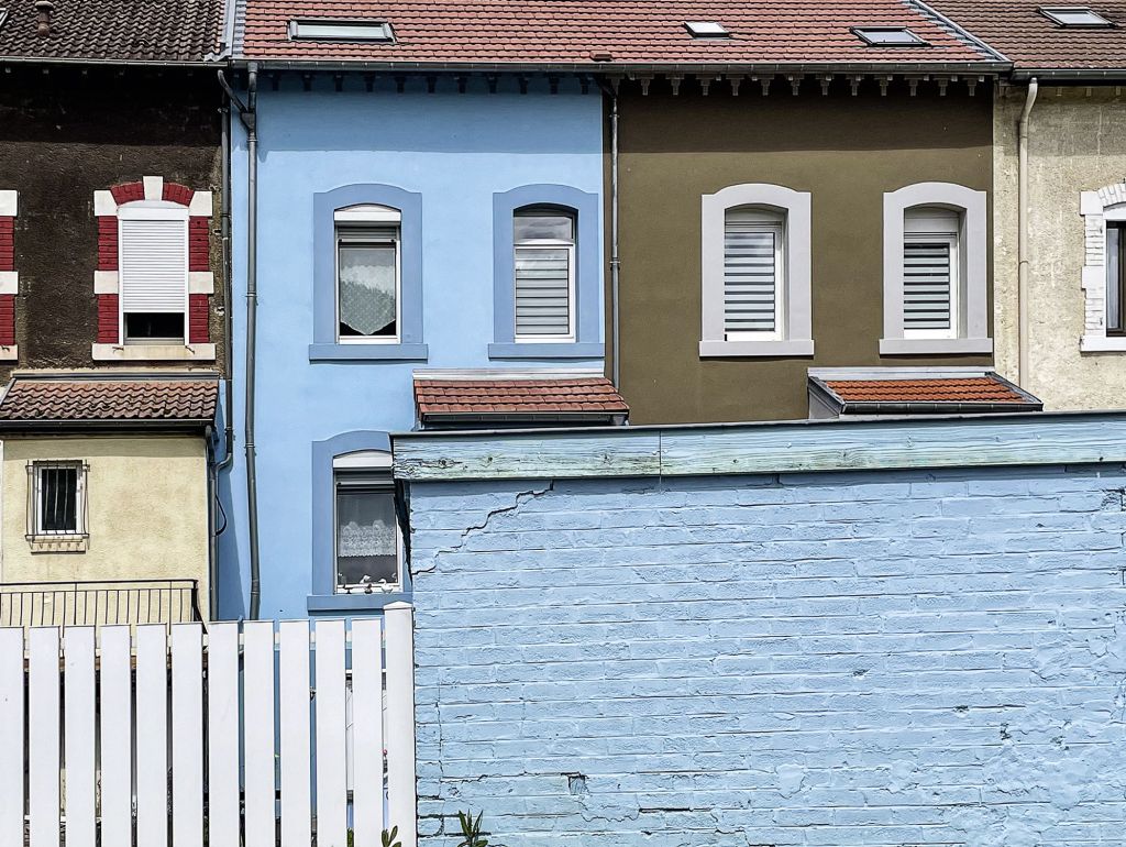 Straßenansicht mit dicht aneinanderstehenden, farbenfrohen Häuserfassaden in Blau, Olivgrün, Beige und Dunkelbraun. Im Vordergrund eine hellblaue, verwitterte Backsteinmauer mit weißem Holzzaun; darüber sind Fenster und rote Ziegeldächer zu sehen.