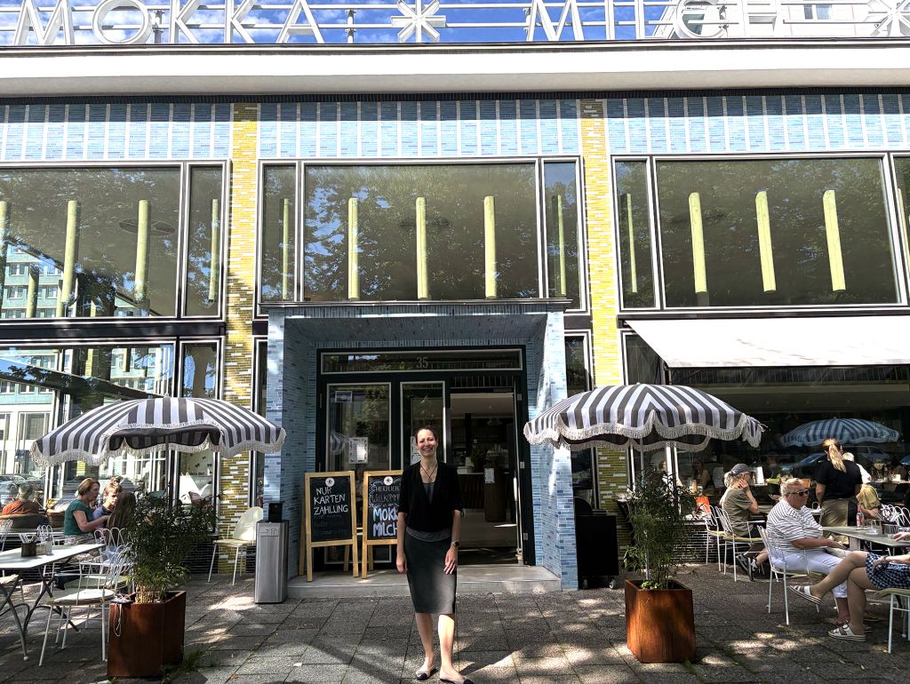 Außenansicht eines Cafés mit großer Glasfront und gelb-weißer Klinkerfassade. Vor dem Eingang stehen zwei gestreifte Sonnenschirme mit Sitzplätzen.  Unternehmerin Natacha Neumannsteht vor der Eingangstür; weitere Gäste sitzen auf der Außenterrasse.