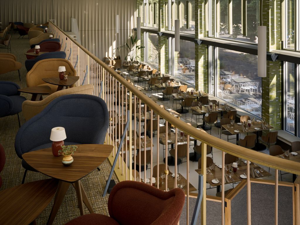 Blick von einer Galerie über einen großzügigen, lichtdurchfluteten Restaurantbereich mit vielen gedeckten Tischen. Eine Holztreppe und ein Geländer führen durch den Raum; bodentiefe Fenster sorgen für viel Tageslicht.