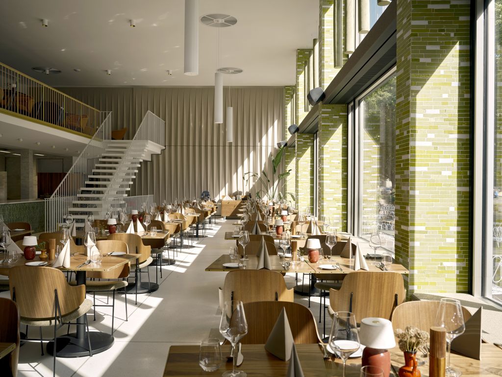 Heller, großzügiger Restaurantbereich mit zahlreichen Holztischen, gedeckten Tischen und einer weißen Treppe zu einer Galerie. Große Fenster mit grünen Klinkerflächen lassen viel Tageslicht in den Raum.