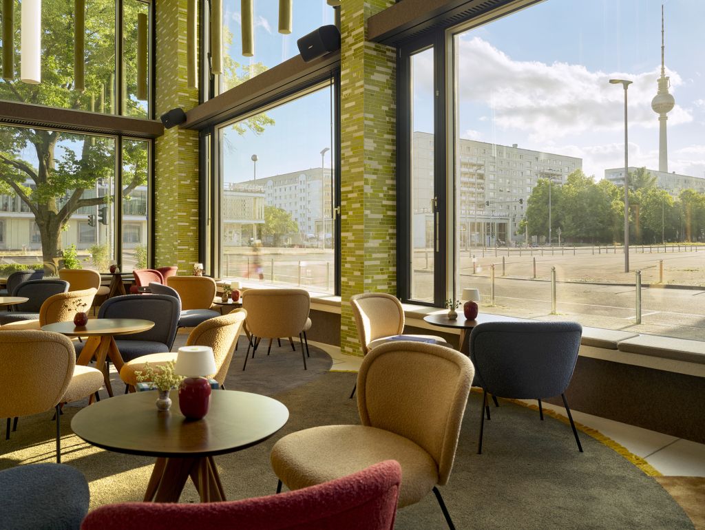 Heller Lounge-Bereich eines Restaurants mit gepolsterten Sesseln und runden Holztischen vor großen Fenstern. Durch die Fenster ist ein weitläufiger Berliner Platz mit Bäumen und Gebäuden zu sehen.
