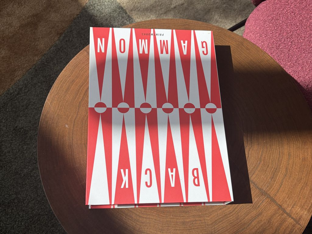 Nahaufnahme eines roten und weißen Back Gammonspiels mit geometrischem Muster auf einem runden Holztisch. Das Magazin liegt in einem hellen Lounge-Bereich mit farbigen Sesseln und Teppichboden.
