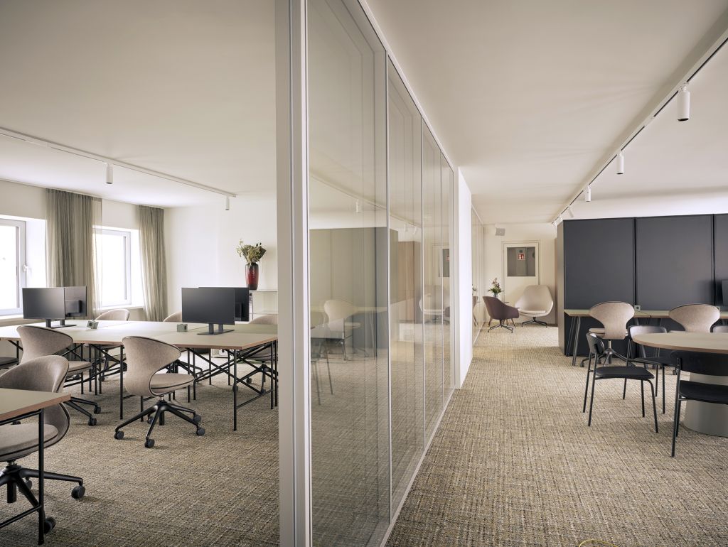 Heller, modern eingerichteter Coworking-Bereich mit Arbeitsplätzen, Computermonitoren und Stühlen. Eine Glaswand trennt den Raum von einem angrenzenden Sitzbereich mit Tischen und Sesseln.