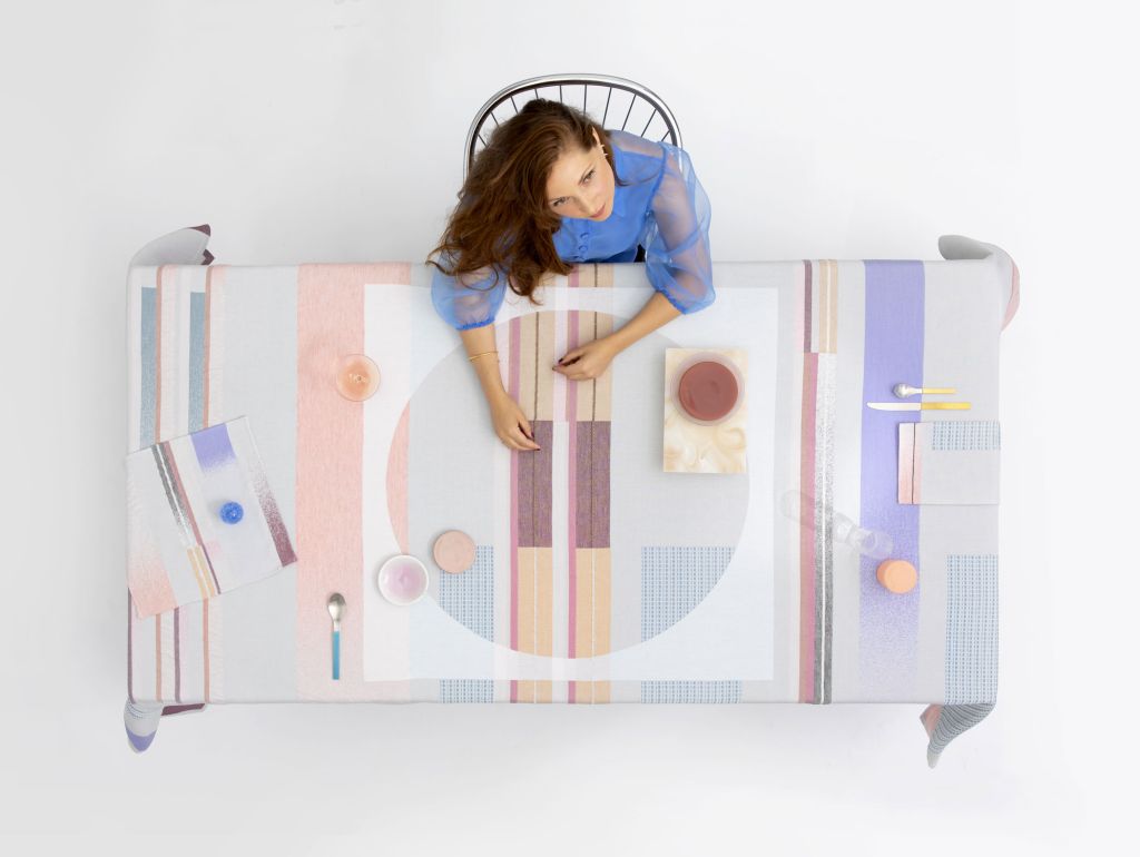 Draufsicht auf eine Frau in einer blauen Bluse, die an einem mit einer pastellfarben gemusterten Tischdecke bedeckten Tisch sitzt. Auf dem Tisch stehen Geschirr, Besteck, Gläser und kleine Schalen mit farbigen Speisen oder Getränken.