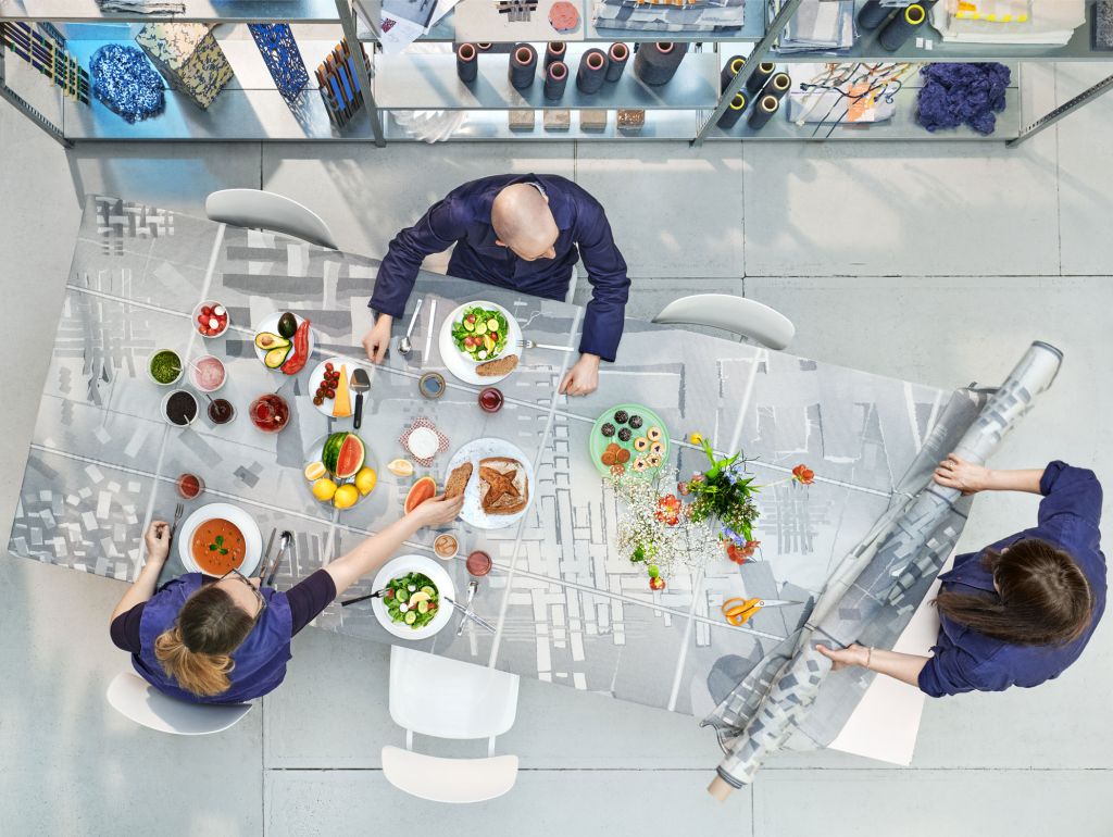Draufsicht auf drei Personen, die an einem großen Tisch mit einer grau gemusterten Tischdecke sitzen. Auf dem Tisch stehen verschiedene Speisen, Obst, Salate, Getränke und Geschirr. Eine Person rechts rollt die Tischdecke teilweise auf; im Hintergrund sind Regale mit Stoffen und Garnen zu sehen.