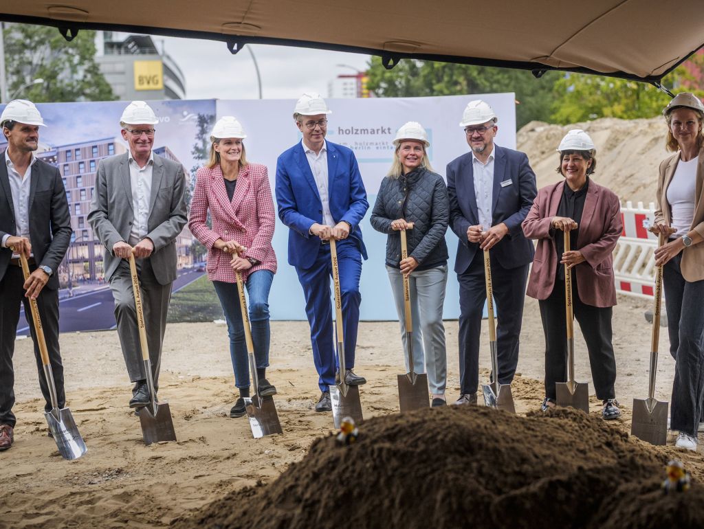 Mehrere Personen mit Schutzhelmen stehen bei einer Baustelle am Holzmarkt mit Spaten vor einem Erdhaufen. Sie präsentieren gemeinsam den offiziellen Spatenstich für ein neues Bauprojekt. Im Hintergrund ist eine Visualisierung des geplanten Gebäudes zu sehen.