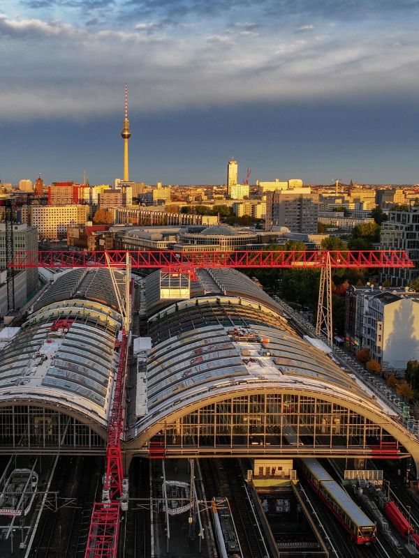 Luftaufnahme des Berliner Ostbahnhofs mit geschwungenen Glasdächern und einem roten Baukran. Im Hintergrund ist der Fernsehturm zu sehen, während die Abendsonne die Gebäude in warmes Licht taucht.