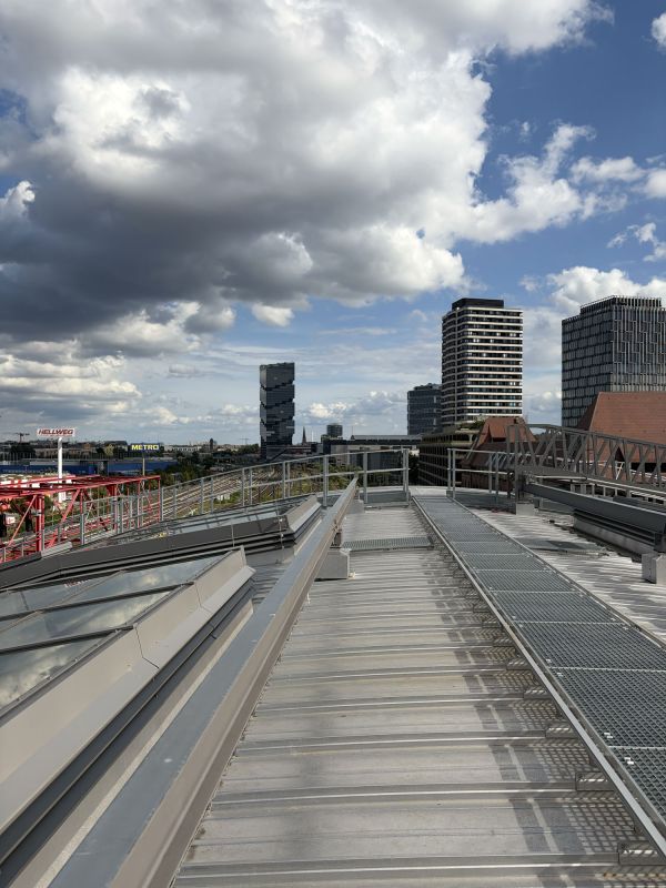 Blick vom Dach des Berliner Ostbahnhofs auf moderne Hochhäuser und die umliegende Stadt. Im Vordergrund sind Metalldachflächen und Glasfenster zu sehen, während große Wolken den blauen Himmel prägen.