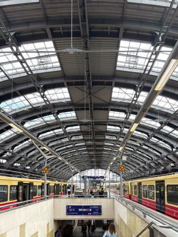 Blick auf einen Bahnhof mit gelben und roten S-Bahn-Zügen unter einer großen, gewölbten Stahl-Glas-Dachkonstruktion. Eine Treppe führt zu den Bahnsteigen hinunter, während Reisende auf den Zug warten.