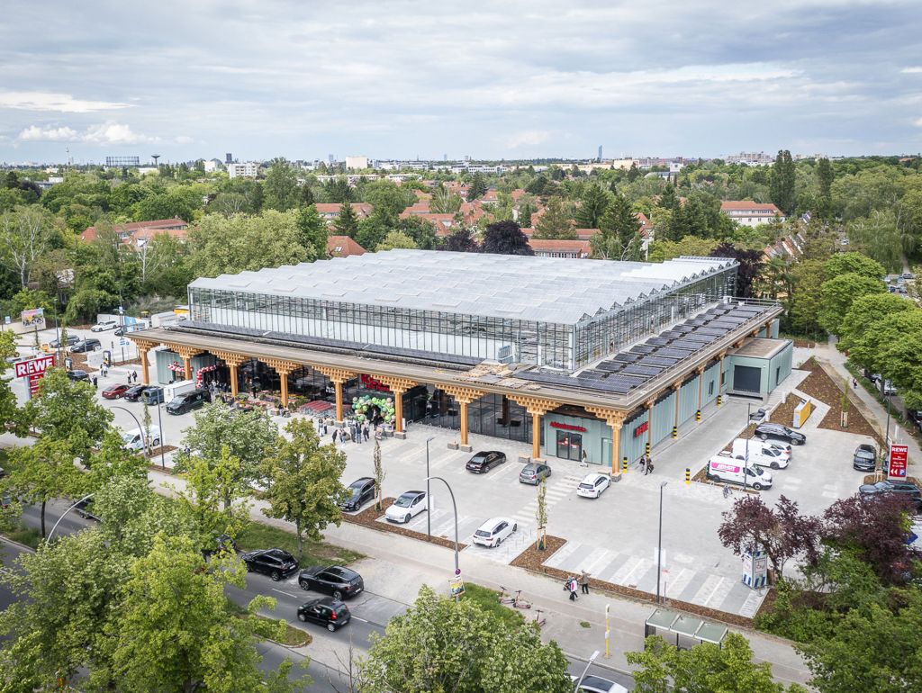 Luftaufnahme eines REWE-Supermarkts mit großem Glasgewächshausdach, Solaranlagen, Parkplatz und umliegenden Bäumen.