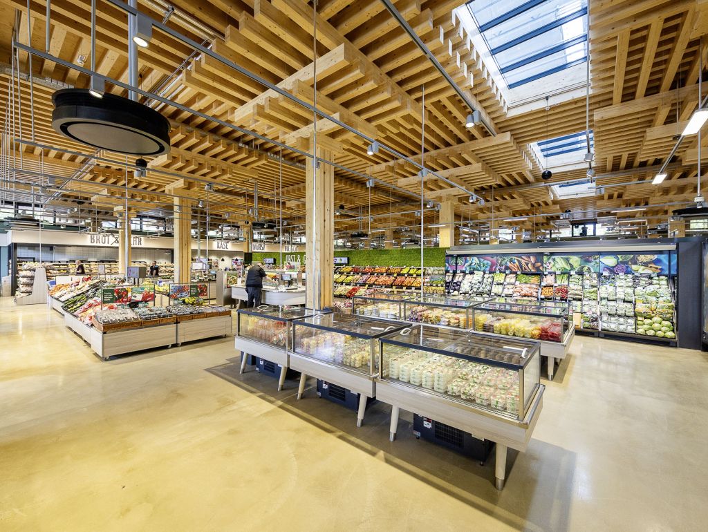  Moderner REWE-Supermarkt mit einer großen Auswahl an frischen Lebensmitteln, Obst und Gemüse sowie gekühlten Produkten. Die offene Holzdeckenkonstruktion und die großen Dachfenster sorgen für eine helle, freundliche Atmosphäre.