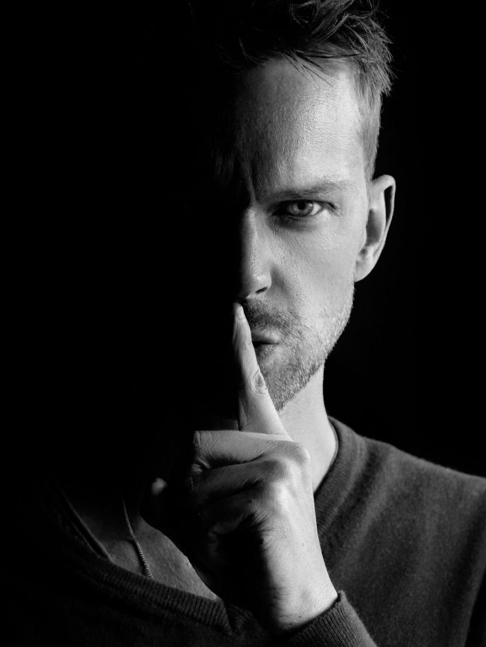 Schwarz-Weiß-Porträt eines Mannes mit kurzem Haar und Bart, der mit einem Finger vor den Lippen eine Geste zum Schweigen macht. Eine Gesichtshälfte liegt im Schatten, der Hintergrund ist schwarz.