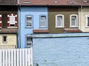 Straßenansicht mit dicht aneinanderstehenden, farbenfrohen Häuserfassaden in Blau, Olivgrün, Beige und Dunkelbraun. Im Vordergrund eine hellblaue, verwitterte Backsteinmauer mit weißem Holzzaun; darüber sind Fenster und rote Ziegeldächer zu sehen.