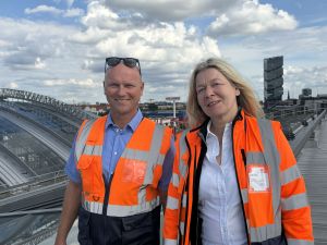 Zwei Personen in orangefarbenen Warnwesten stehen auf dem Dach des Berliner Ostbahnhofs. Im Hintergrund sind die Glasdachkonstruktion und die Stadtkulisse zu sehen.